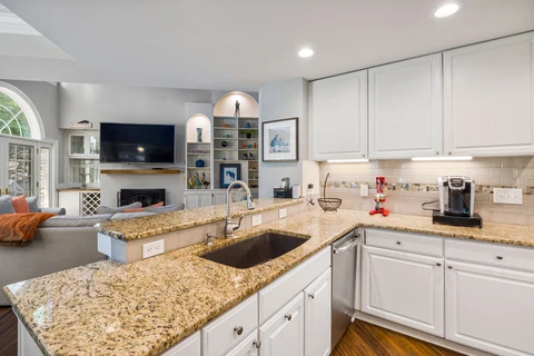 A kitchen with white cabinets and granite counters opens onto a living area.