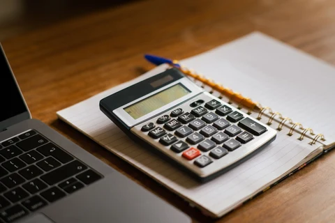 A calculator rests on an open notebook beside a laptop on a wooden table.