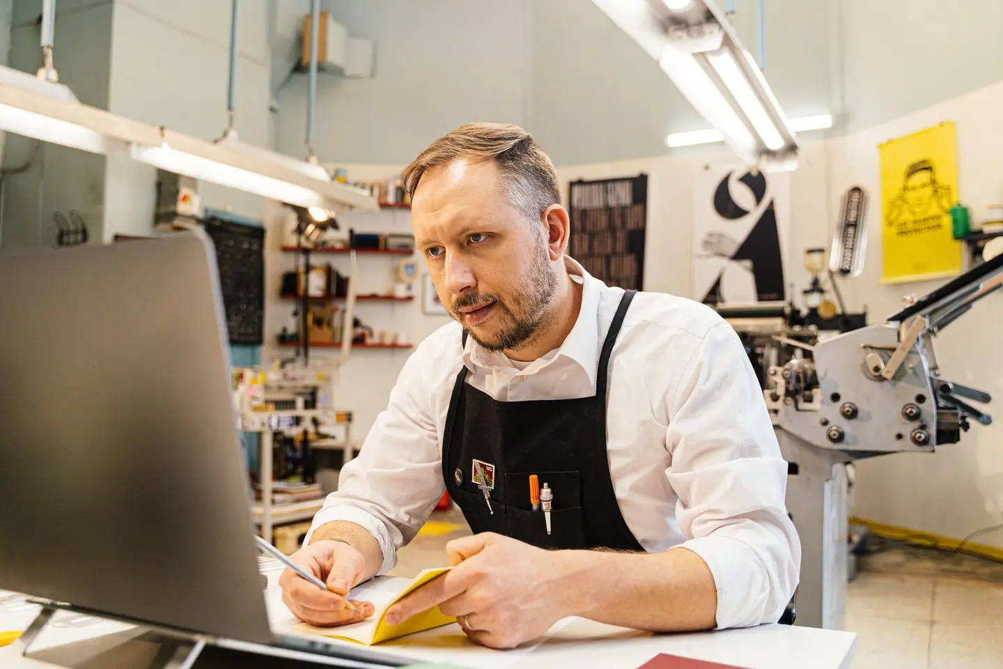 A man in an apron works at a laptop beside printing equipment.
