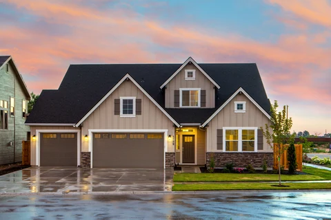 A two-story house with an illuminated entry and garage beside a wet street at sunset.