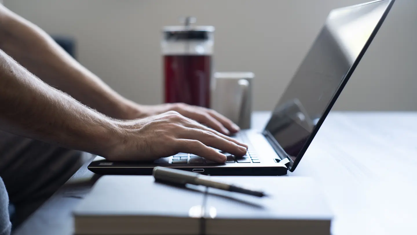 Hands typing on a laptop beside a notebook