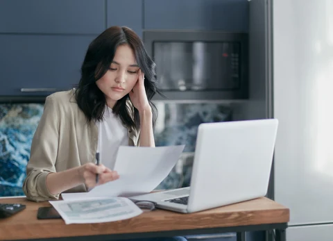 A woman reviews paperwork beside an open laptop at a table.